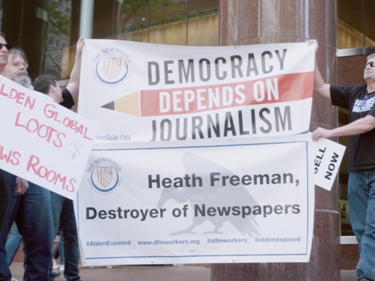 A still image from Stripped From Parts of journalists protesting. Two hold up a large sign with "Democracy Depends on Journalism" and "Heath Freeman, Destroyer of Newspapers."